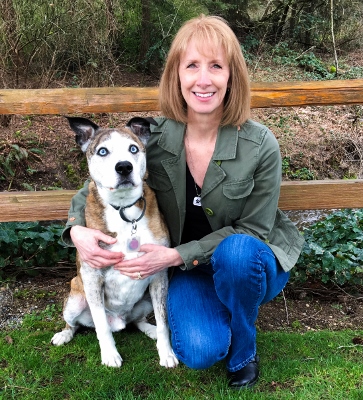 A woman with shoulder-length light brown hair kneels on grass next to a brown and white dog with blue eyes. They are outdoors by a wooden fence with trees and greenery in the background.