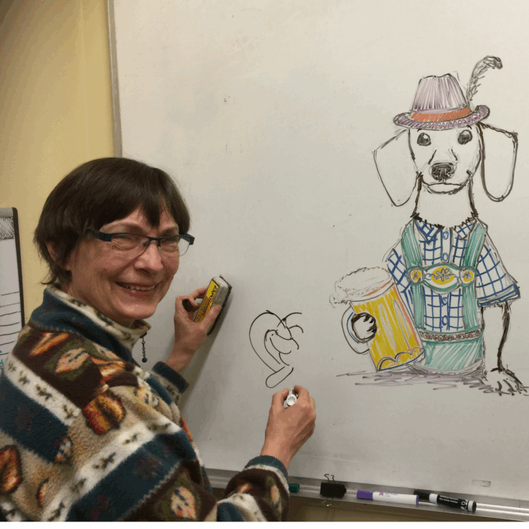 A woman smiles while drawing on a whiteboard. Next to her is a detailed drawing of a dog wearing traditional Bavarian clothes, holding a beer mug, and wearing a feathered hat.