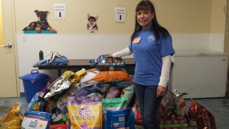A woman in a blue shirt stands smiling beside a large pile of donated pet food bags inside a room decorated with posters of dogs and labeled donation signs on the wall behind her.