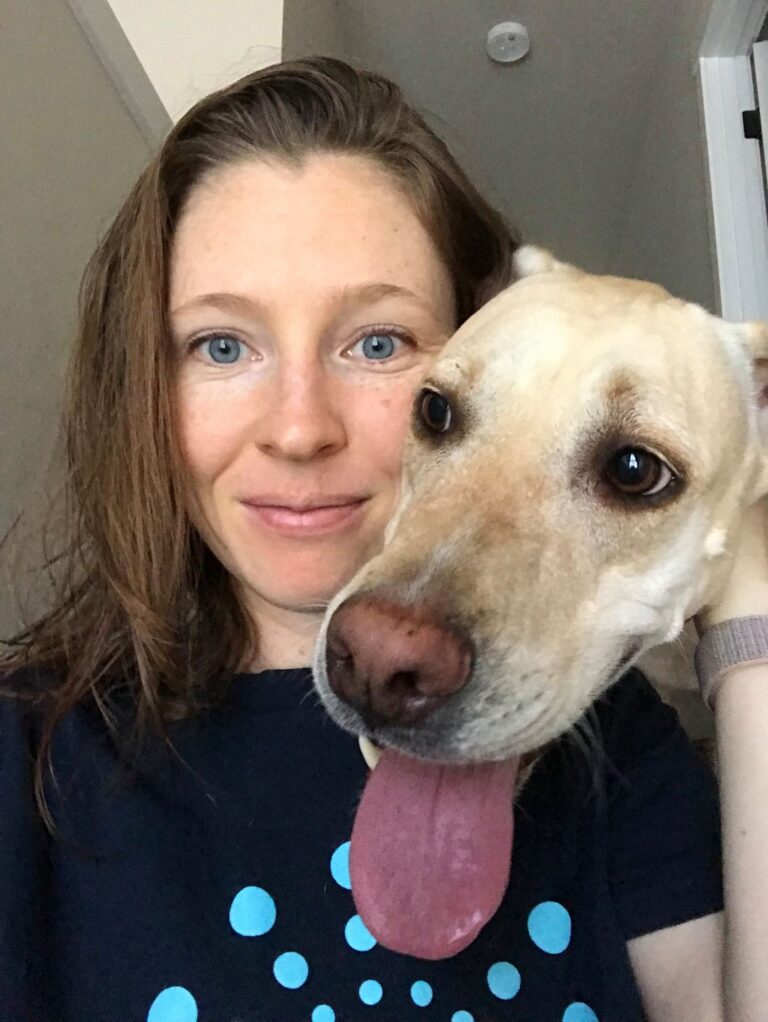 A woman with light brown hair smiles at the camera, while a tan dog with large brown eyes and its tongue out leans in close beside her. They appear to be indoors near a white door.