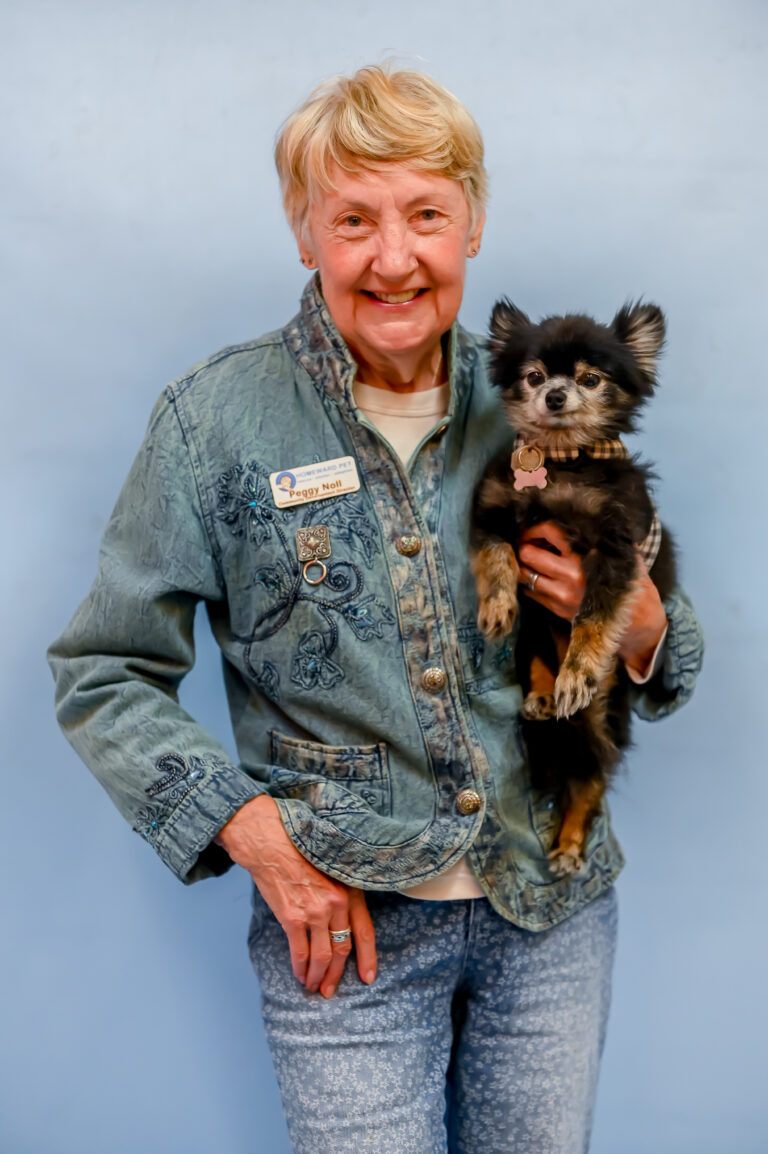 An older woman with short blonde hair wearing a blue patterned jacket and jeans smiles while holding a small black and brown dog. She has a name tag on her jacket and stands against a plain light blue background.