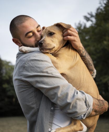 A man with a beard and tattoos hugs and kisses a tan dog outdoors, both appearing content and affectionate, with trees and sky in the background.