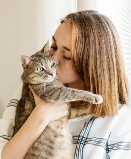 A woman with shoulder-length light brown hair holds and kisses a gray tabby cat near a window. The cat looks slightly to the side while the woman affectionately embraces it.