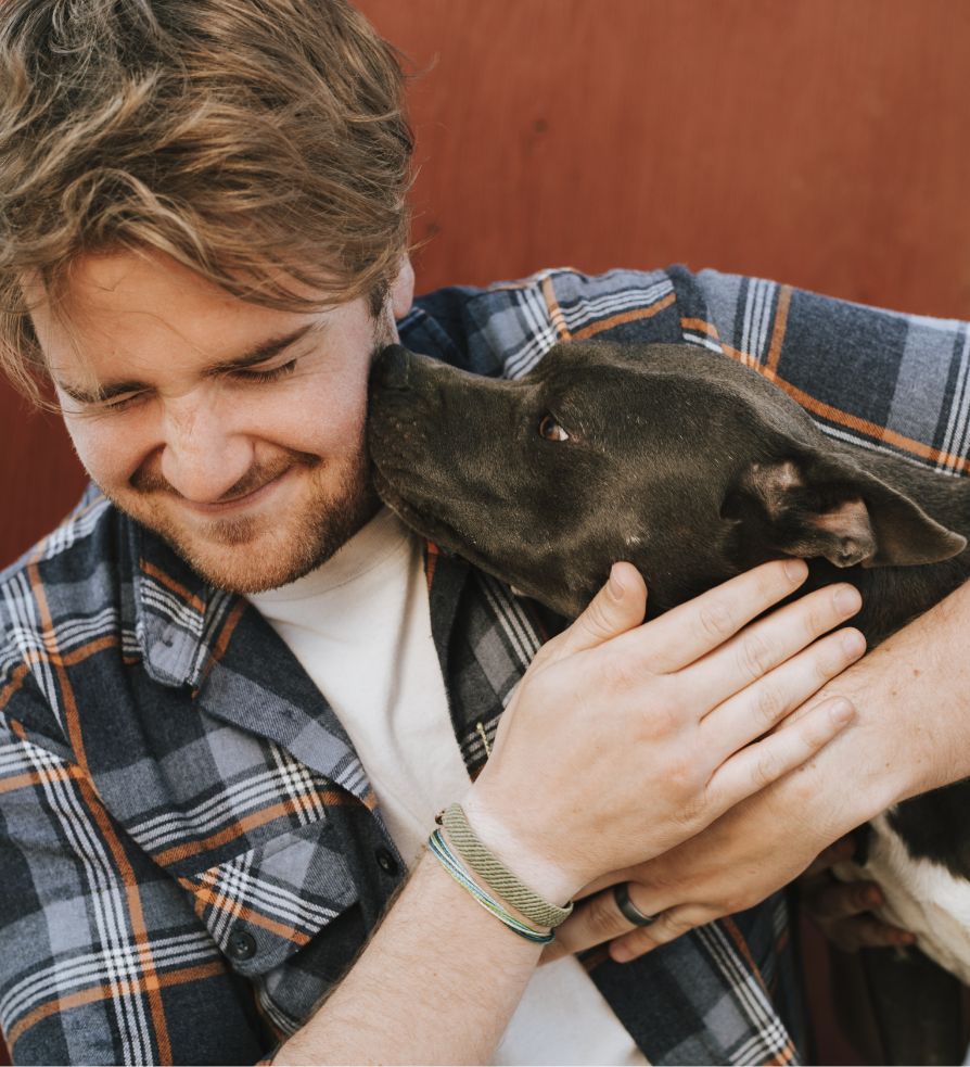 A man with brown hair smiles with his eyes closed as a black and white dog affectionately licks his cheek. The man hugs the dog and wears a plaid shirt, while the background is a reddish wall.