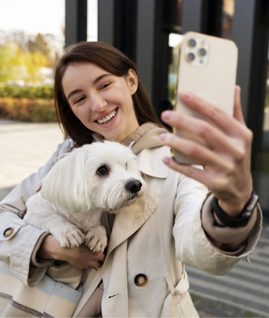 A young woman with brown hair smiles while taking a selfie outdoors, holding a small white dog in her arms. Both appear happy, and the background includes blurred greenery and modern buildings.