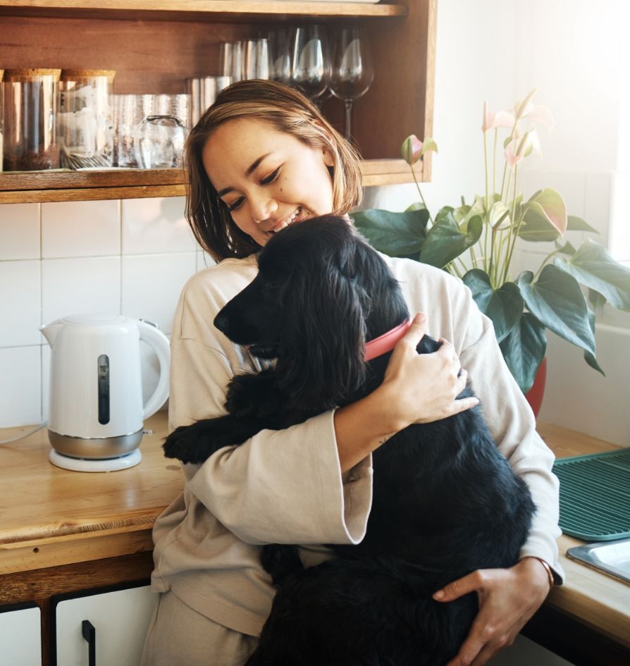 A woman in light-colored clothes smiles while hugging a black dog in a kitchen. There is a kettle, glasses, and a potted plant on the counter beside her. Sunlight streams in through a window.