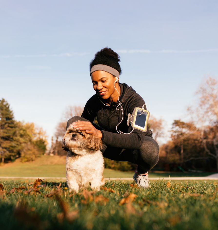 A woman in athletic wear, with earbuds and a smartphone armband, kneels on grass and pets a small, fluffy dog. Trees and blue sky are in the background, suggesting a park setting on a sunny day.