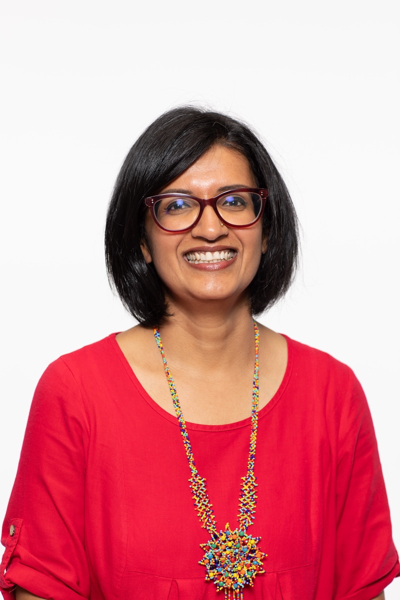 A woman with short black hair and glasses smiles at the camera. She is wearing a bright red top and a large, colorful beaded necklace. The background is plain white.