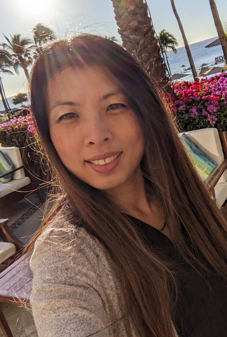 A woman with long brown hair takes a selfie outdoors in sunny weather. She smiles slightly, with palm trees, colorful flowers, patio furniture, and an ocean view with a clear sky in the background. Sunlight creates a lens flare.