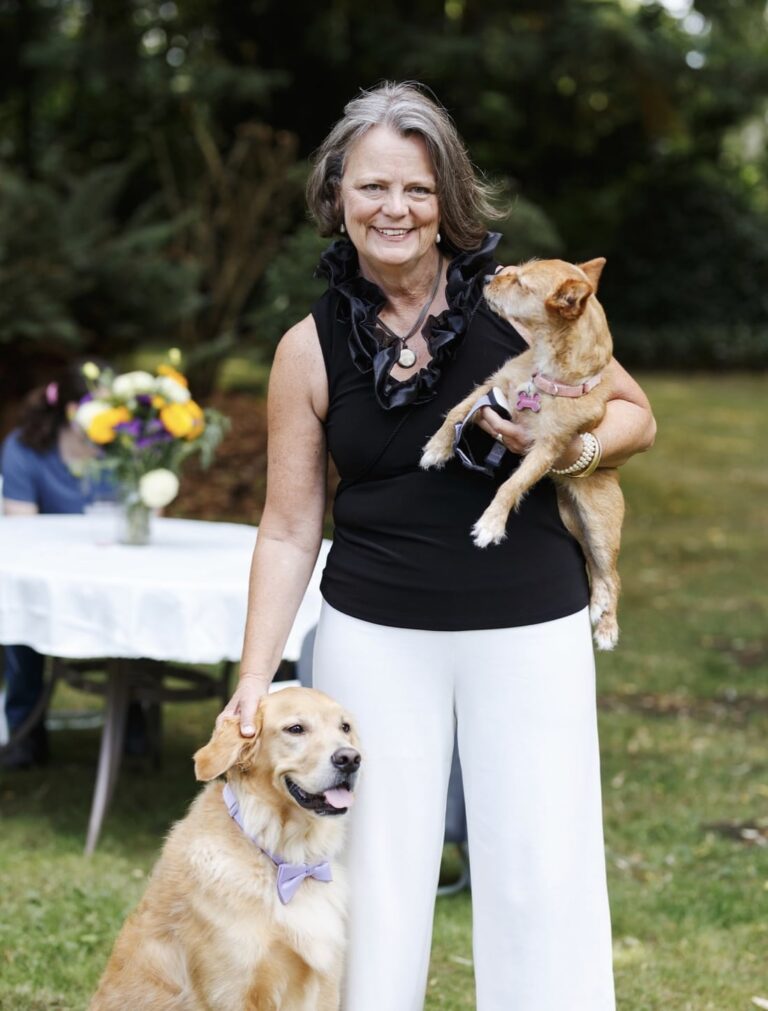 A smiling woman in a black sleeveless top and white pants stands outdoors, holding a small brown dog and petting a golden retriever. A table with flowers and greenery is visible in the background.