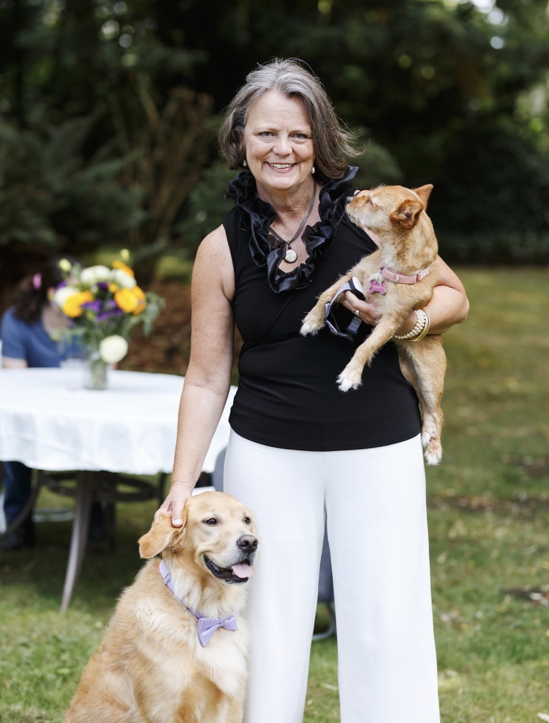 A smiling woman in a black sleeveless top and white pants stands outdoors, holding a small brown dog and petting a golden retriever. A table with flowers and greenery is visible in the background.
