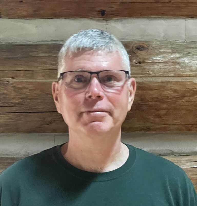 A man with short gray hair, glasses, and a dark green shirt stands in front of a wooden log wall, looking straight at the camera with a neutral expression.