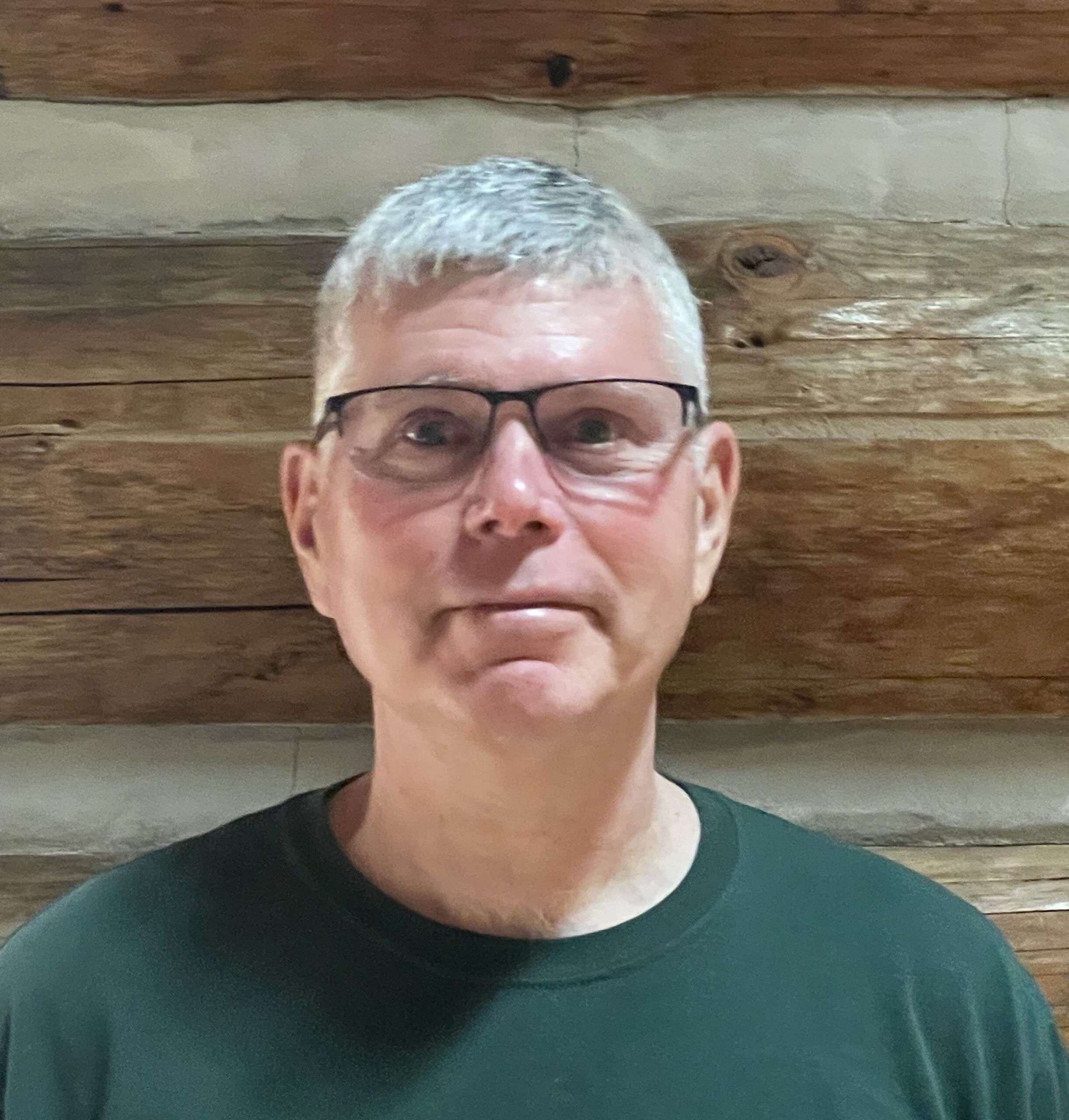 A man with short gray hair, glasses, and a dark green shirt stands in front of a wooden log wall, looking straight at the camera with a neutral expression.